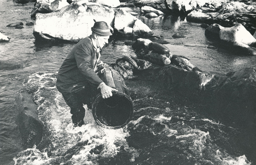 svartvitt fotografi av stövelförsedd man som står i älv med en spolformad, tunnliknande tina avsedd att fånga fisken med.