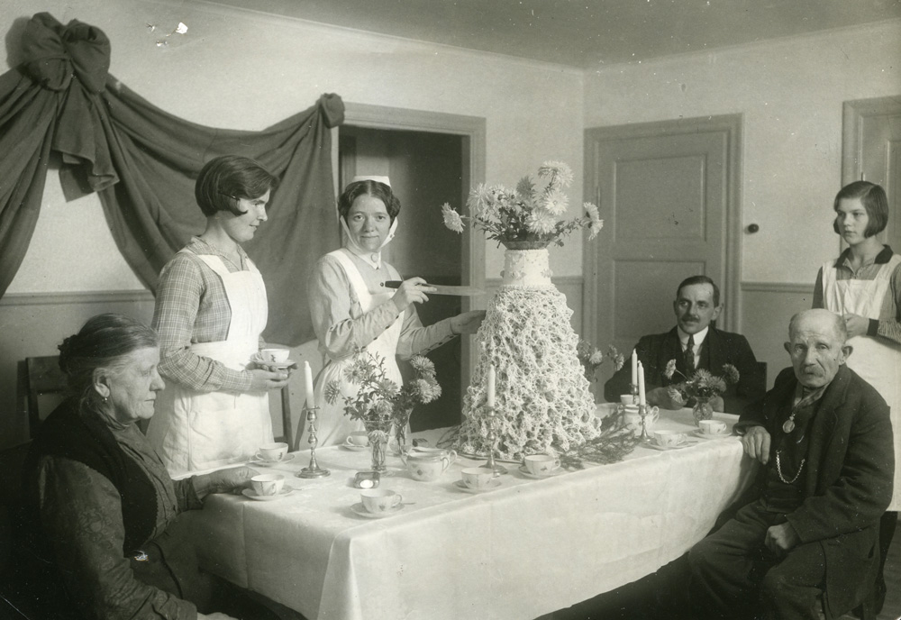 Svartvitt fotografi. Människor runt ett kalasdukat bord med en hög spettekaka i mitten. En sköterskeklädd kvinna håller på att skära upp kakan.